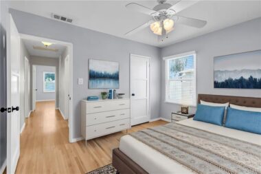 Digitally staged back bedroom with ceiling fan, hardwood floors, new window blinds, double pane windows, and view into the hallway.