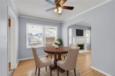 Digitally staged photo of formal dining room with hardwood floors, new window blinds, ceiling fan, and view into the living room.